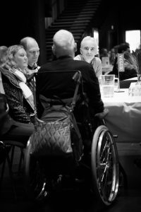 En åben bog om handicap sidder i kørestol ved et bord omgivet af lånere der stiller spørgsmål.