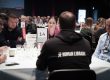 A Human Library book with a group of readers sitting at a table with many other tables also full at a conference hall.