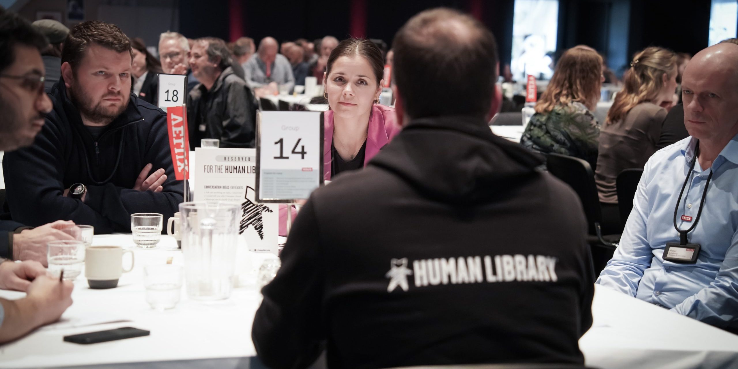 A Human Library book with a group of readers sitting at a table with many other tables also full at a conference hall.