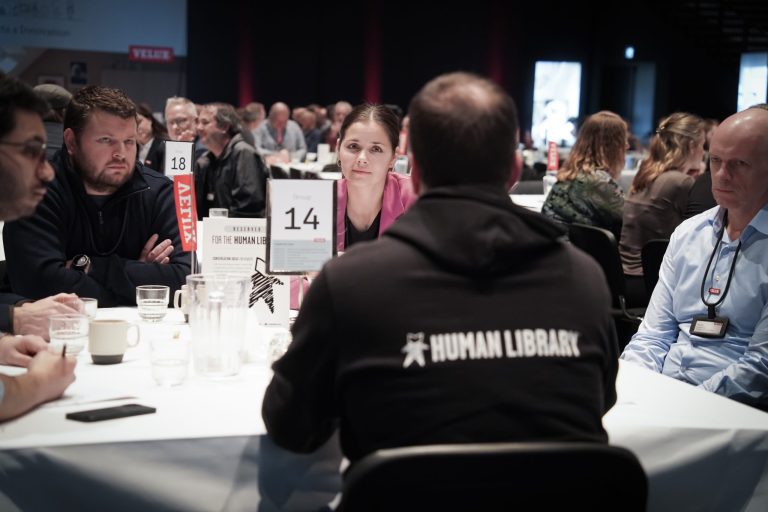 A Human Library book with a group of readers sitting at a table with many other tables also full at a conference hall.