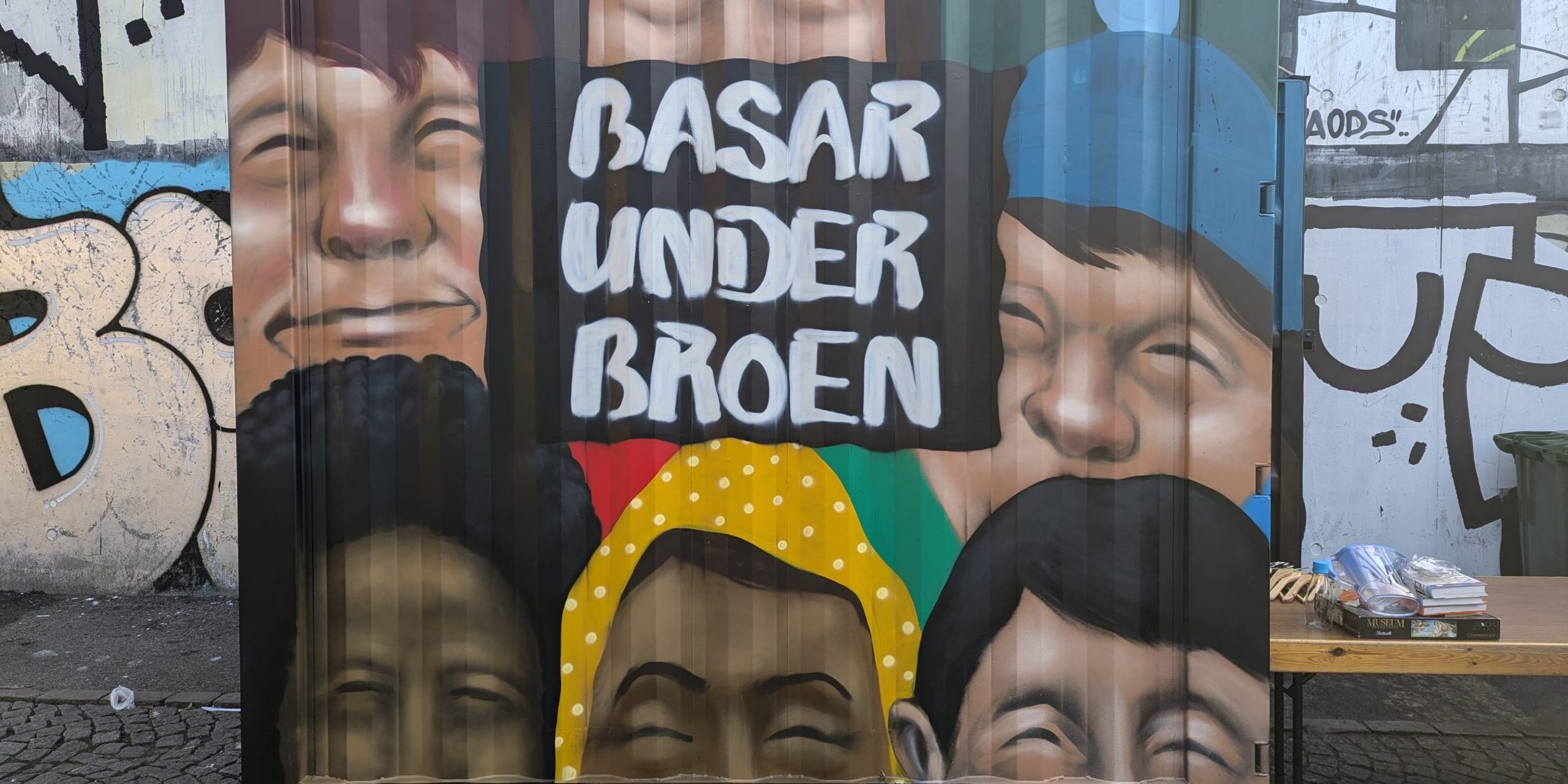 Container som opbevarer borde og tøjstativer for basaren under broen malet med graffitti seks ansigter og navnet på basaren.