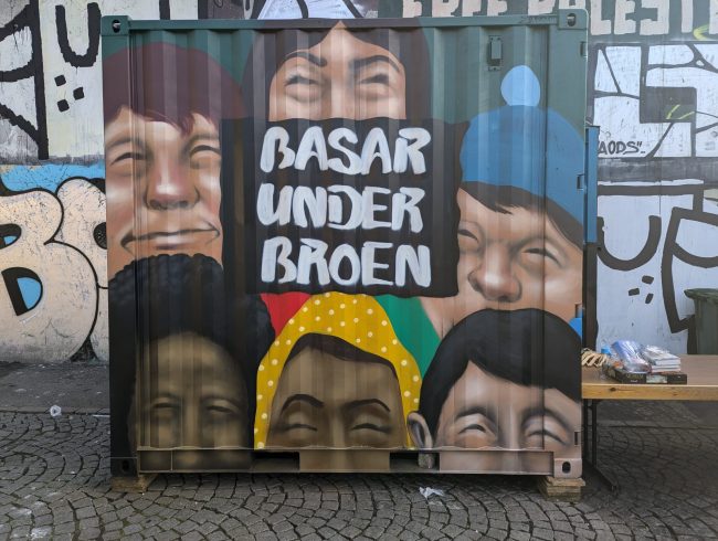 Container som opbevarer borde og tøjstativer for basaren under broen malet med graffitti seks ansigter og navnet på basaren.