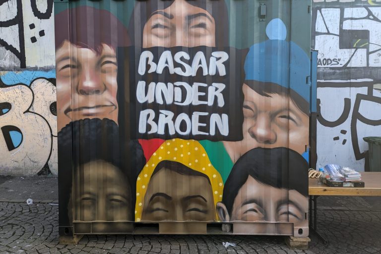 Container som opbevarer borde og tøjstativer for basaren under broen malet med graffitti seks ansigter og navnet på basaren.